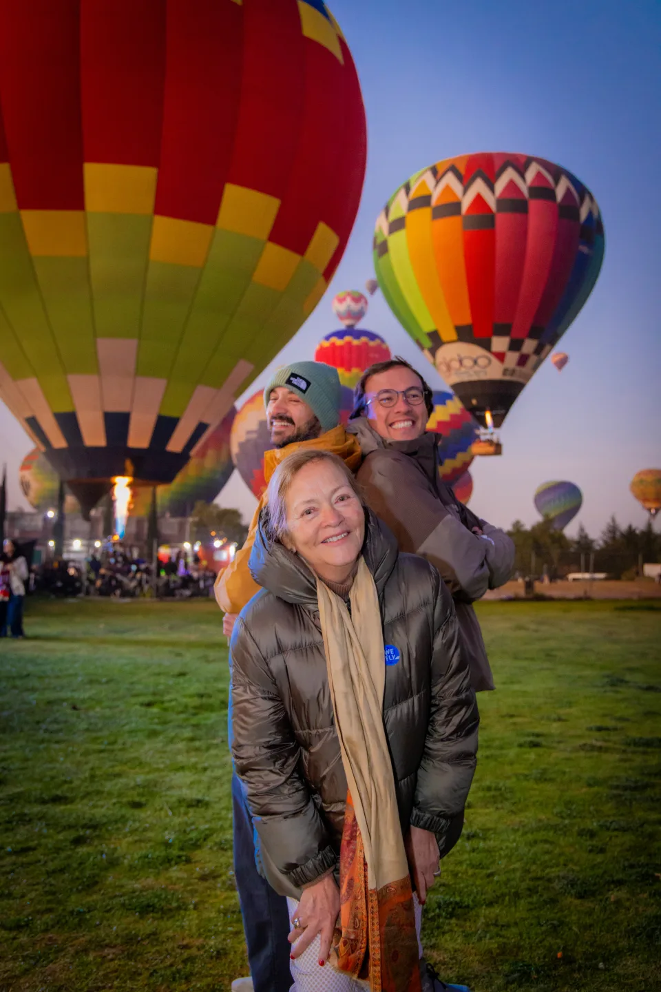 Madre cargando a sus dos hijos adultos en la espalda, riendo ante globos encendidos al amanecer en Teotihuacán
