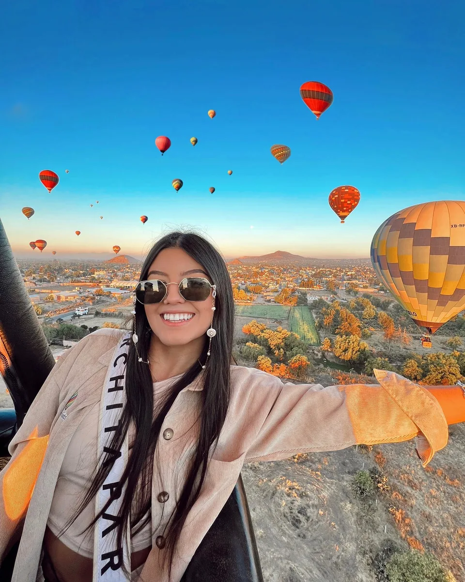 A passenger opens both arms in the basket as the sky fills with balloons at sunrise