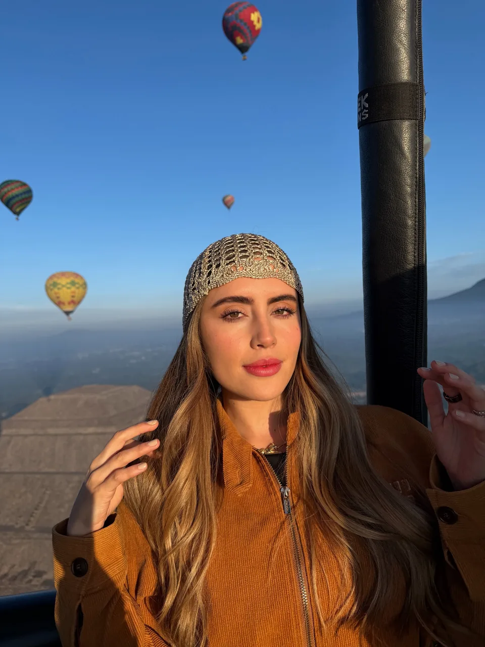 A passenger beside the burner frame with balloons rising over the valley behind