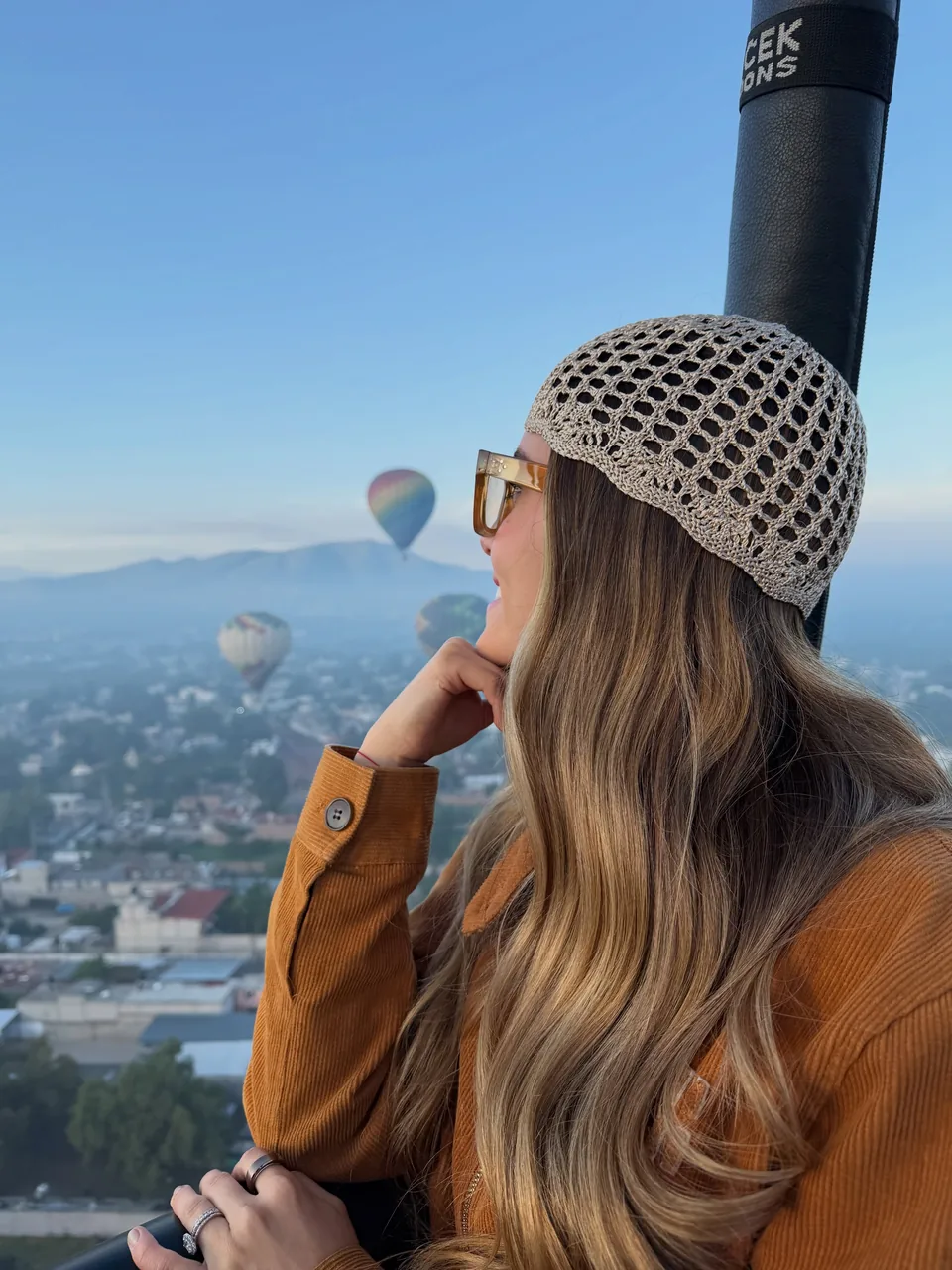 A passenger resting on the basket rail with balloons scattered over the valley behind