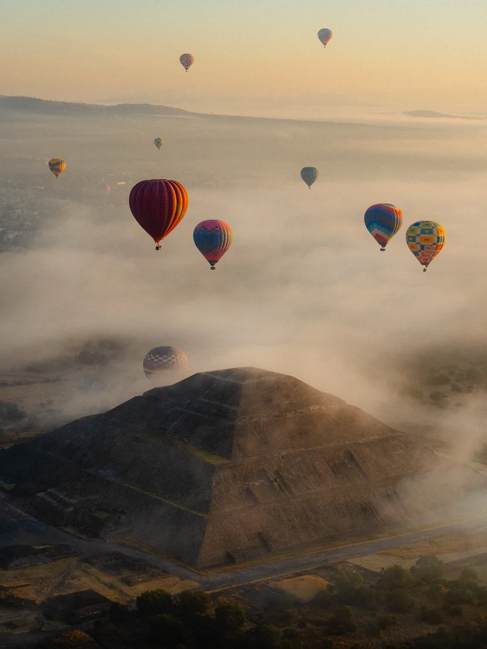 A pyramid at Teotihuacan half covered in low morning fog, with balloons floating above it