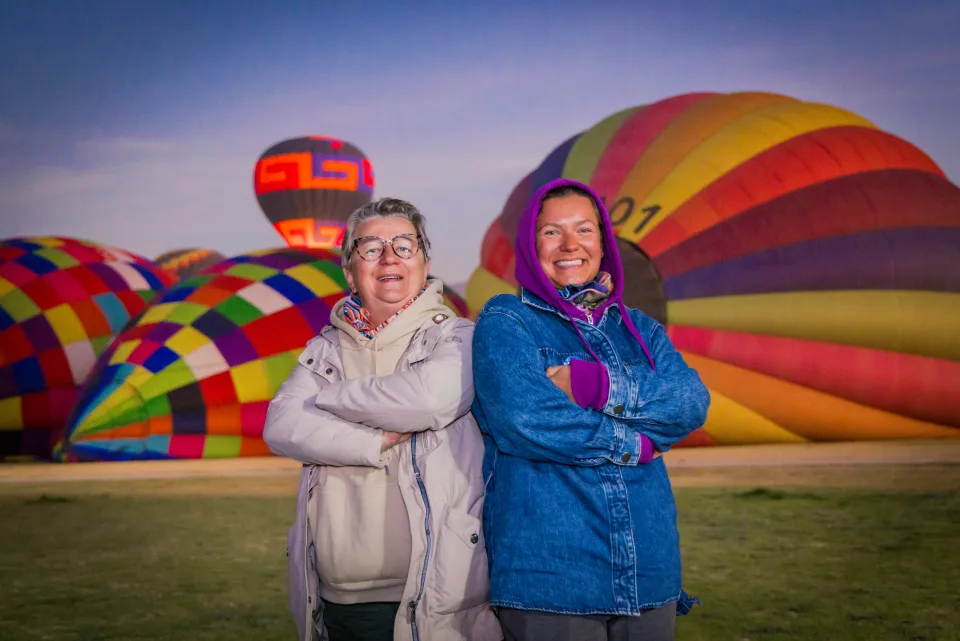 Dos mujeres posando espalda con espalda con coloridos globos inflándose al amanecer