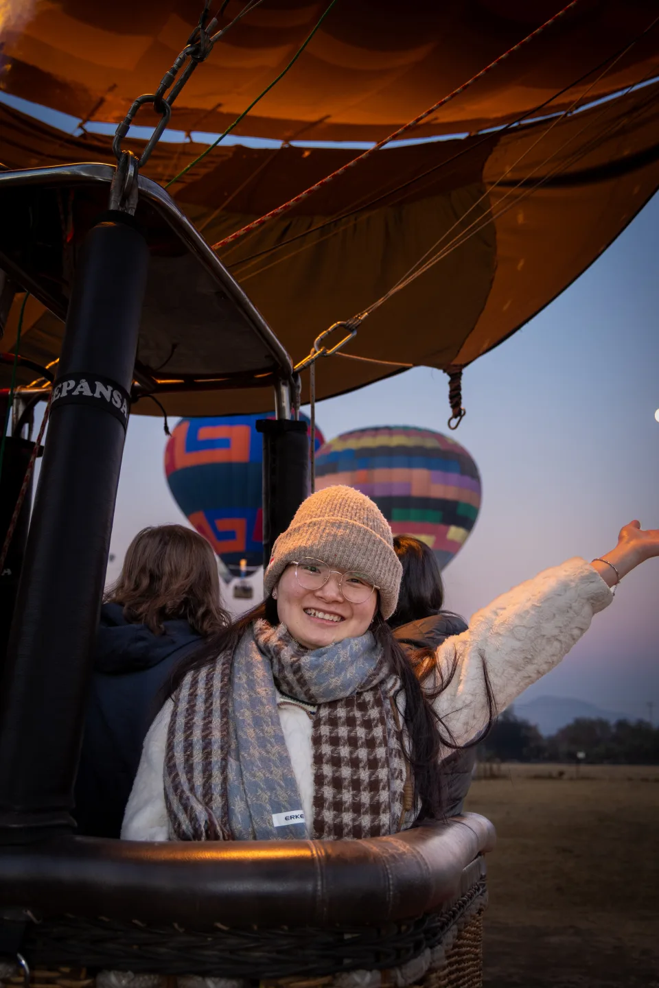 Mujer abrigada sonriendo en la canastilla con globos flotando detrás al amanecer